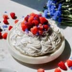A pavlova topped with whipped cream, puree, raspberries and cornflower on the counter.