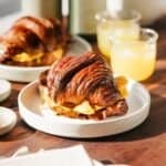 Bacon egg and cheese croissant on a plate with juice nearby on a counter.
